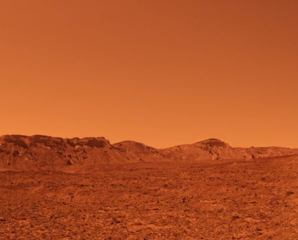 A landscape scene of Mars. It is desert-like and sandy with mountains in the background. The air is hazy and the whole image is tinted red.