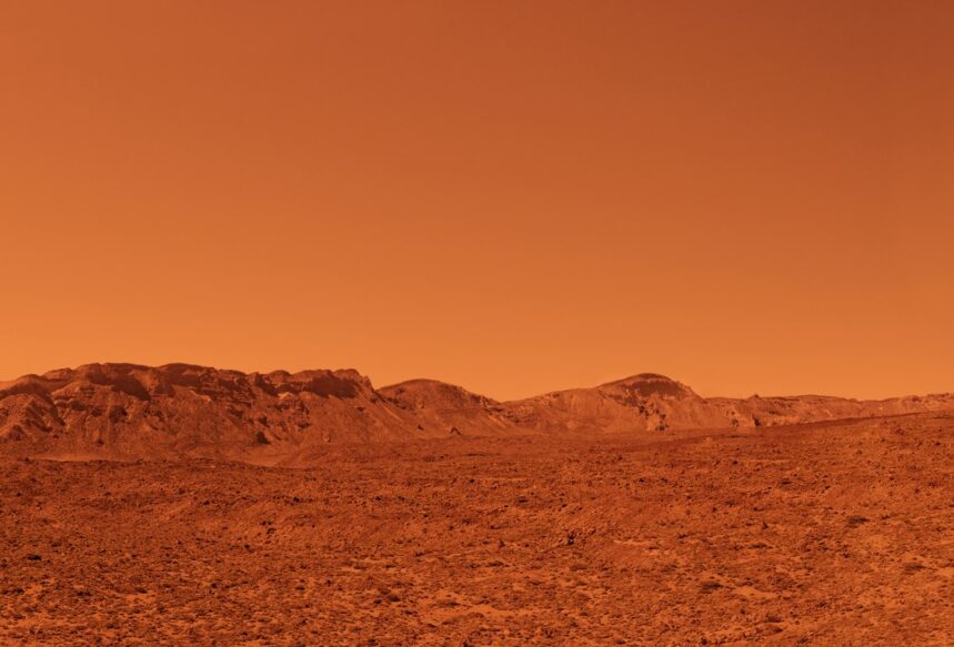 A landscape scene of Mars. It is desert-like and sandy with mountains in the background. The air is hazy and the whole image is tinted red.