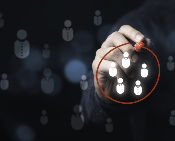 A hand reaching out from a dark background holding a red marker. They are using the marker to draw a red circle around a group of white icons representing people. Other faded icons of people appear in the background.