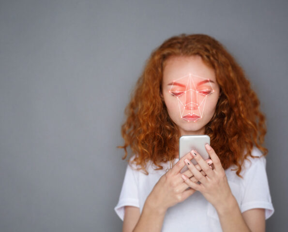 Young woman holding her phone in front of her face against a grey background. There is a red, face-scanning laser grid on her face.