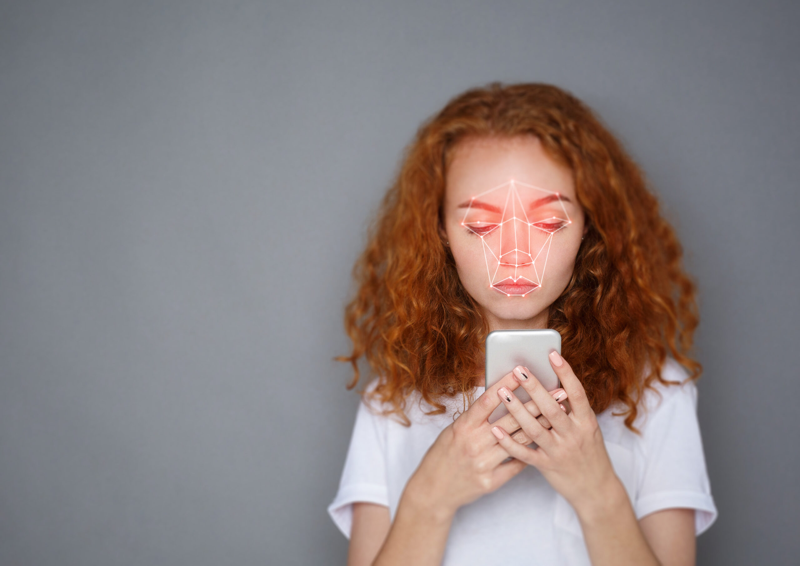 Young woman holding her phone in front of her face against a grey background. There is a red, face-scanning laser grid on her face.