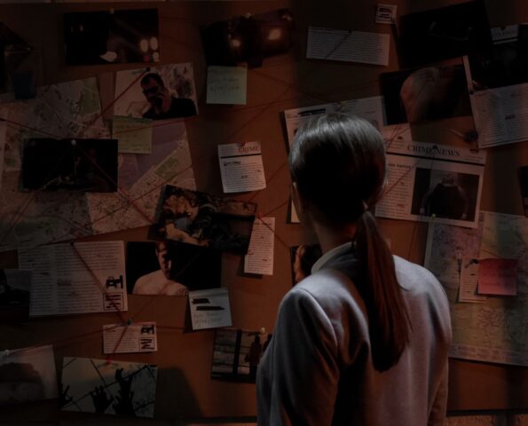 Dark photo of a female detective looking at a cork board. On the board there are notes and newspaper clippings.