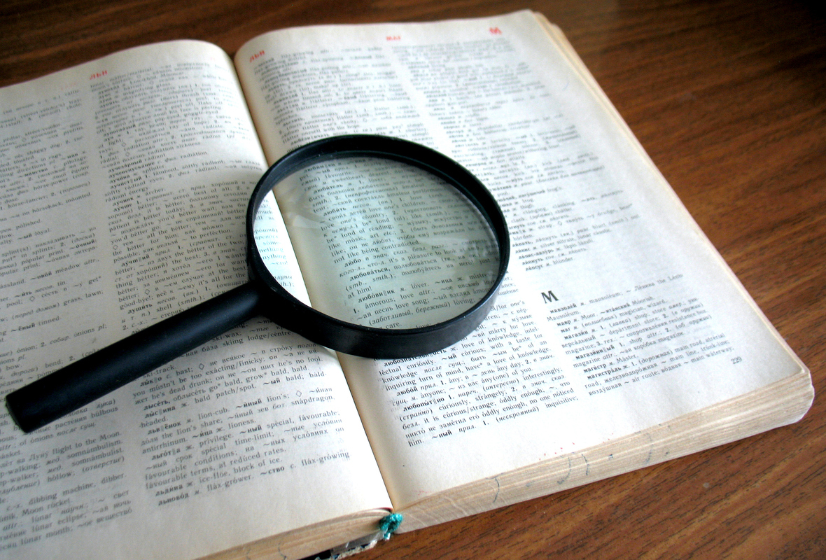 Photograph of an open dictionary on a desk. On top of the pages is a magnifying glass.
