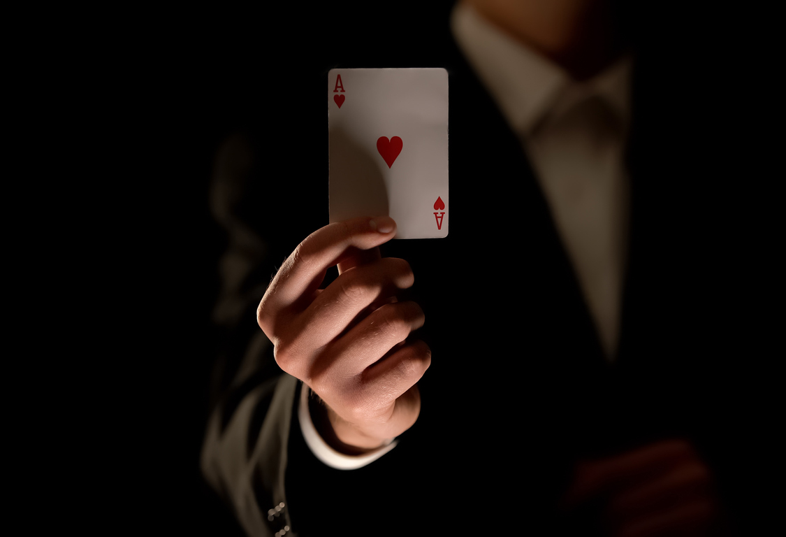 A man in a suit against a dark background holding a playing card up towards the camera. It is the ace of hearts.